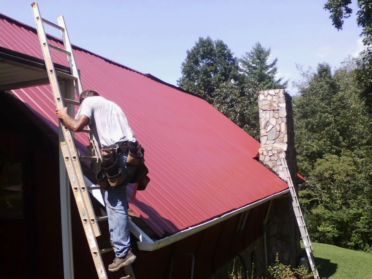 Roofer on a ladder beside a red metal roof and stone chimney — Soffit & Fascia Repair by Roofing Brothers in Tyrone